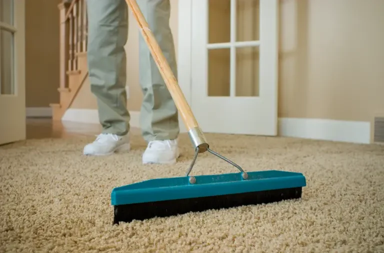 Person using a carpet rake to lift carpet fibres and loosen dirt before deep cleaning with carpet cleaner shampoo.
